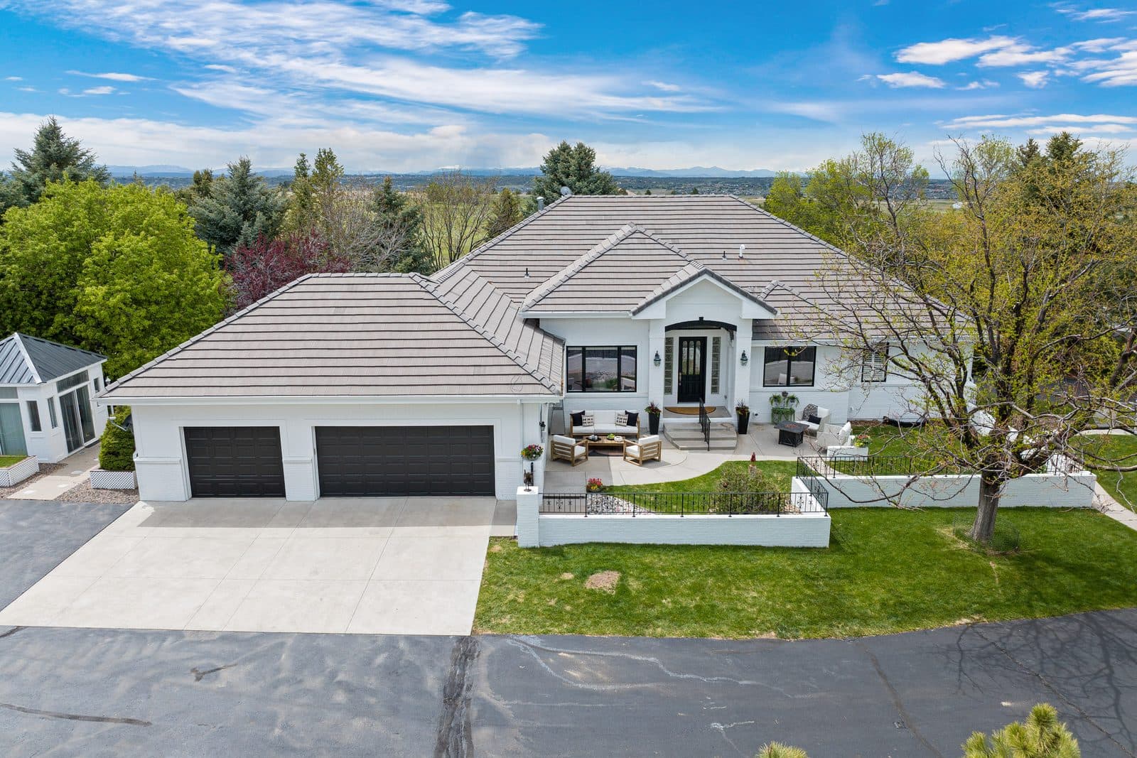 A Douglas County property from the air: a white single-story house with a tiled hip roof and three-car garage, low white garden walls around a lawn, and the Front Range along the horizon.