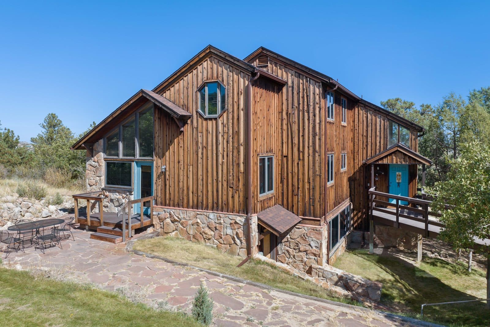 A Douglas County house clad in weathered vertical cedar over a rough stone base, with a steep gabled roof, turquoise door and window frames and dry native grass running out to ponderosa pines.