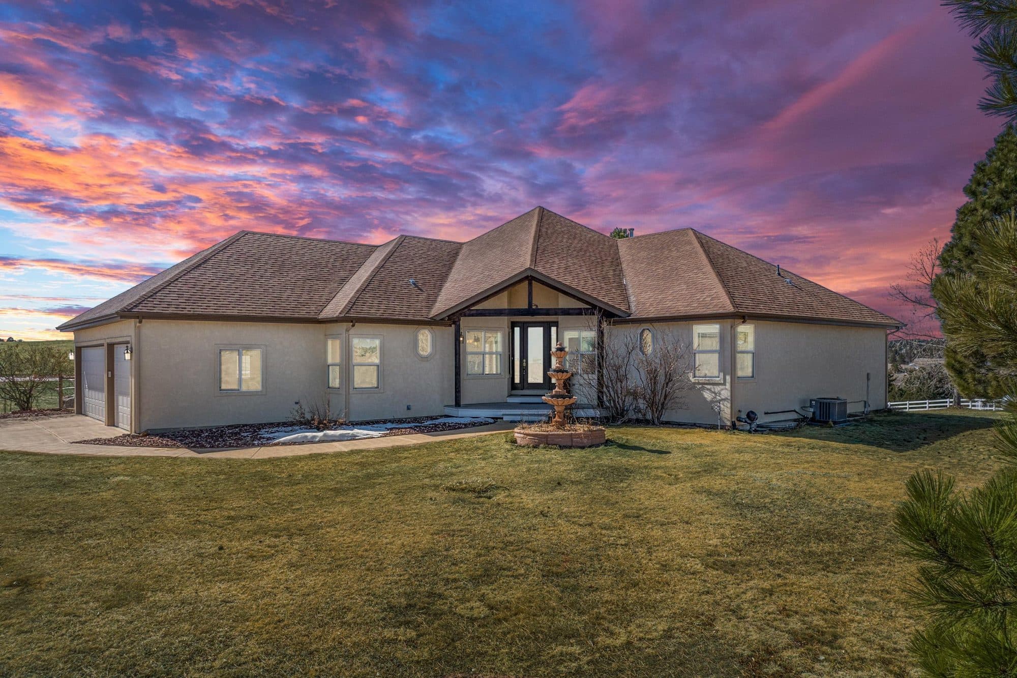 An Elbert County ranch home at dusk under a pink and violet sky: a long stucco single-story house with a hipped roof, a lit entry and a stone fountain on the lawn.